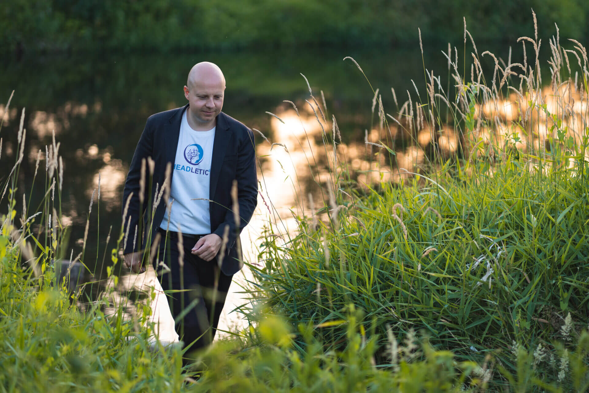 Ein Mann geht bei Sonnenuntergang inmitten von hohem Gras an einem Fluss entlang und trägt ein T-Shirt mit Headletic Logo.