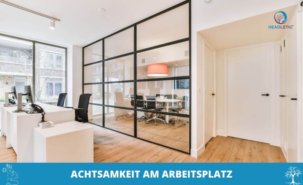 Modernes Büro mit Schreibtischen und Stühlen im Vordergrund, einem gläsernen Besprechungsraum mit rundem Tisch und Stühlen sowie einem Schild, das auf die Bedeutung von Achtsamkeit am Arbeitsplatz hinweist.