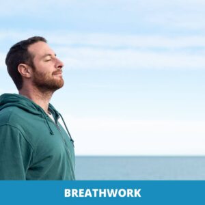 Ein Mann in einem grünen Kapuzenpulli steht mit geschlossenen Augen am Meer und übt Atemarbeit, während er unter einem klaren Himmel tief einatmet. Am unteren Rand steht das Wort "BREATHWORK".