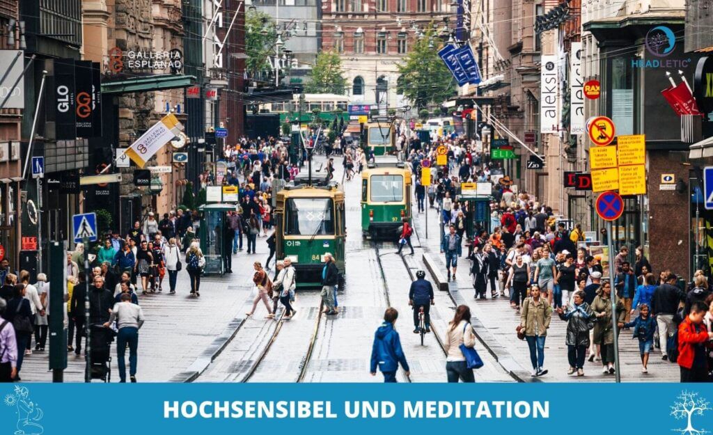 Eine belebte Straße mit Straßenbahnen, Geschäften und Menschenmassen; leuchtende Schilder umrahmen die lebhafte Szene. Der Text am unteren Rand lautet "HOCHSENSIBEL MEDITATION" und hebt einen achtsamen Ansatz für sensible Menschen inmitten des städtischen Chaos hervor.
