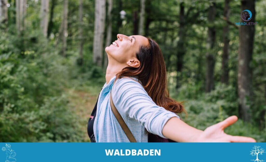 Eine Frau steht auf einem Waldweg mit ausgestreckten Armen und zurückgelegtem Kopf und genießt die Ruhe Waldbadens zwischen hohen Bäumen. Auf dem blauen Banner unten steht "WALDBADEN.